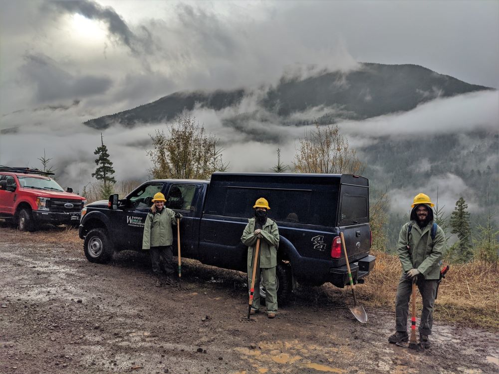 Three WCC members wearing rain gear and hard hats hold tools while standing in front of a WCC truck and smiling at the camera. Behind the truck is forested mountain surrounded by low clouds.