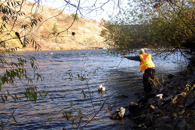 Scientist tosses a group of sample bottles on a rope into a wide river from an Eastern Washington river bank.