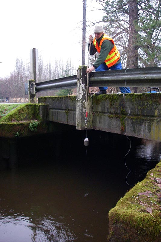Scientist stands on low bridge and lowers sampling bottle down by rope.