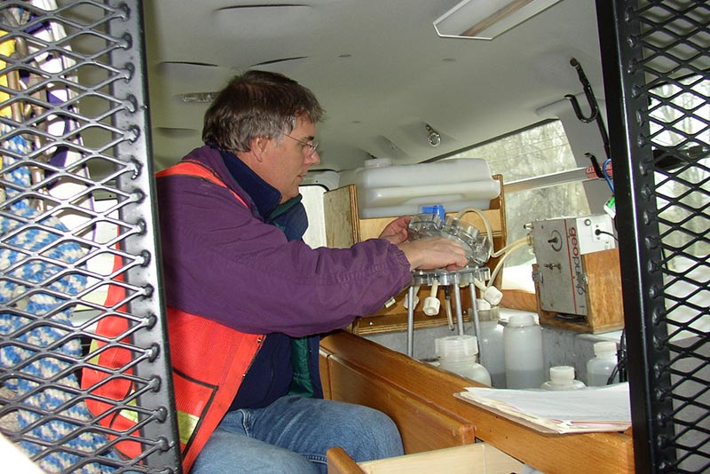 Scientist works with bottles and pump in a laboratory set up inside a sampling van. There is an iron grate in the foreground.