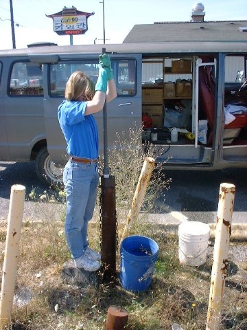 Scientist pulls water from a monitoring well that looks like a rusty pipe in the ground. She is beside a van in a parking lot.
