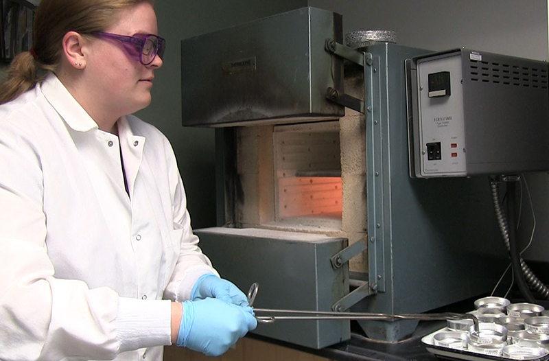 Scientist in a lab coat and purple-rimmed safety goggles uses long tongs to pick up sample crucibles to place in fire-brick lined oven.