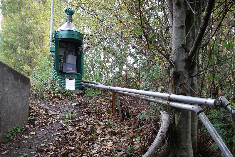 Telemetry station above a stream in a green, metal galvanized  shelter with water pipes protruding. It has a door; the door is open, and equipment is inside. 