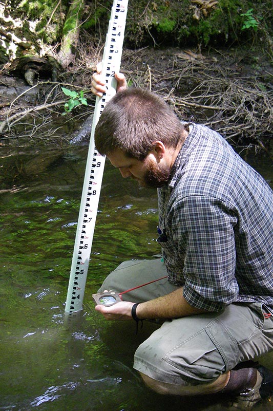 Scientist squats in creek with long white measuring pole marked with incremental numbers and holds a stream gauge.