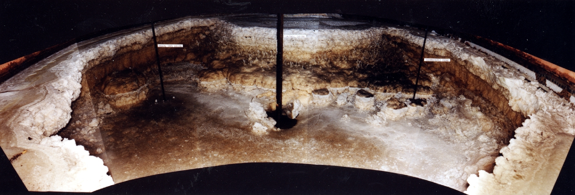 Photo inside an underground storage tank, showing large built up sections of white saltcake around the sides, with the middle full of a dark brown and white specked radioactive sludge.