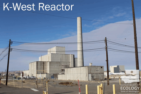 A photo from the ground of Hanford's K-West reactor, a huge concrete building with a tall smoke stack/next is D Reactor, cocooned reactor, large concrete with white roof.