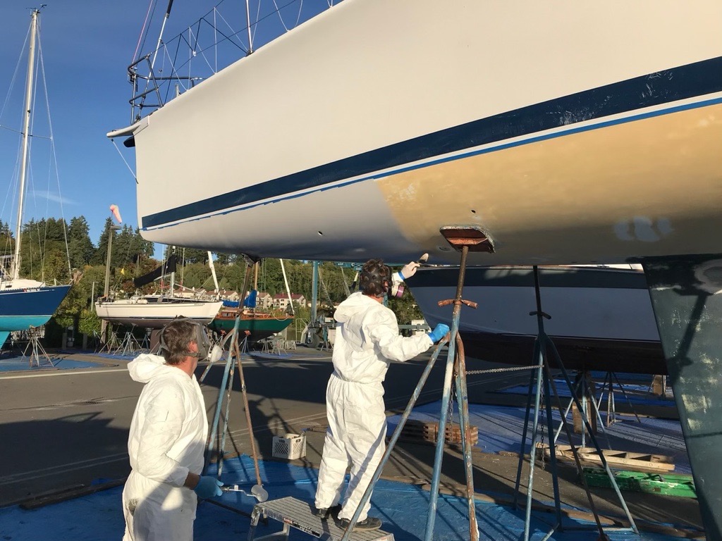 Two people wear respirators and plastic suits to paint a boat hull