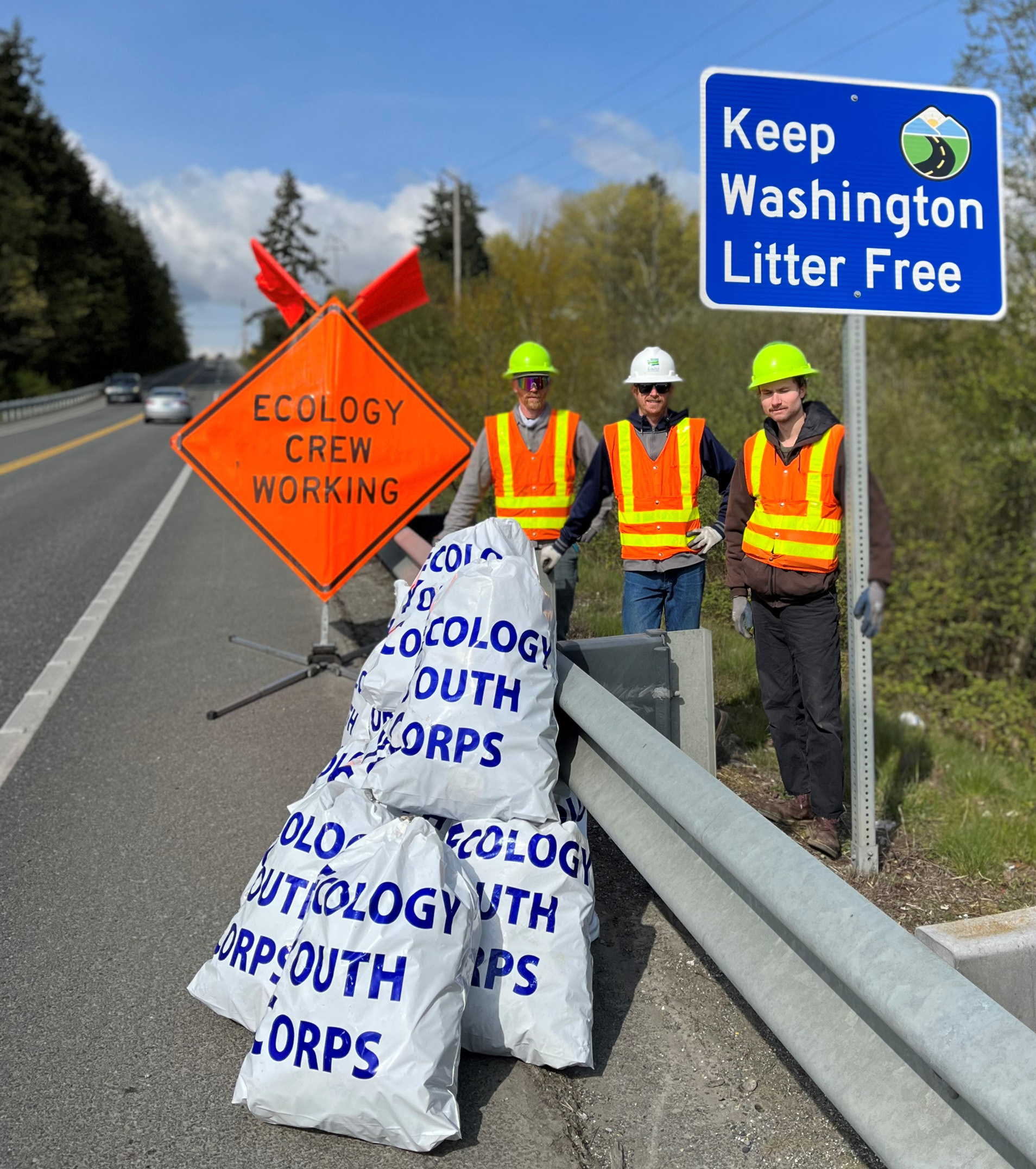 An Ecology Youth Corps litter crew works by a highway, alongside a pile of full litter bags and a new "Keep Washington Litter Free" sign.