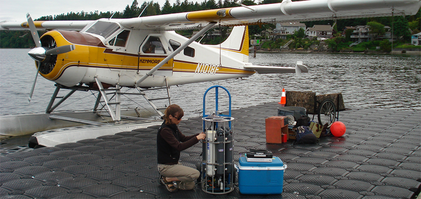 Scientist crouches in front of brown, yellow, and white float plane. She adjusts the equipment in front of her that looks like metal tanks in a cylindrical cage. A blue cooler chest sits nearby.