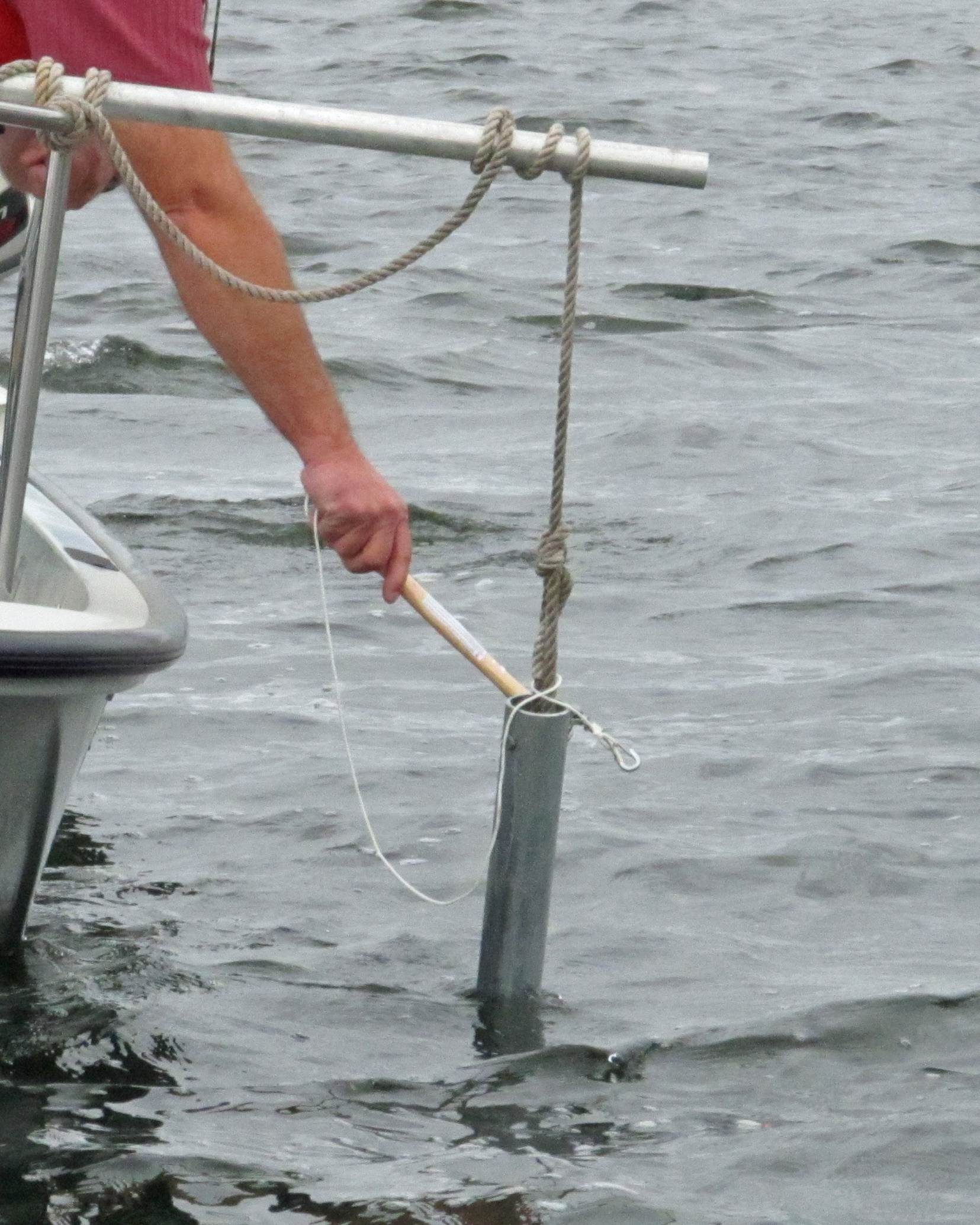 A pipe suspended by cord from a stick is held partly in the water off the side of a small boat, a person's hand holds a metal bar to strike the pipe.