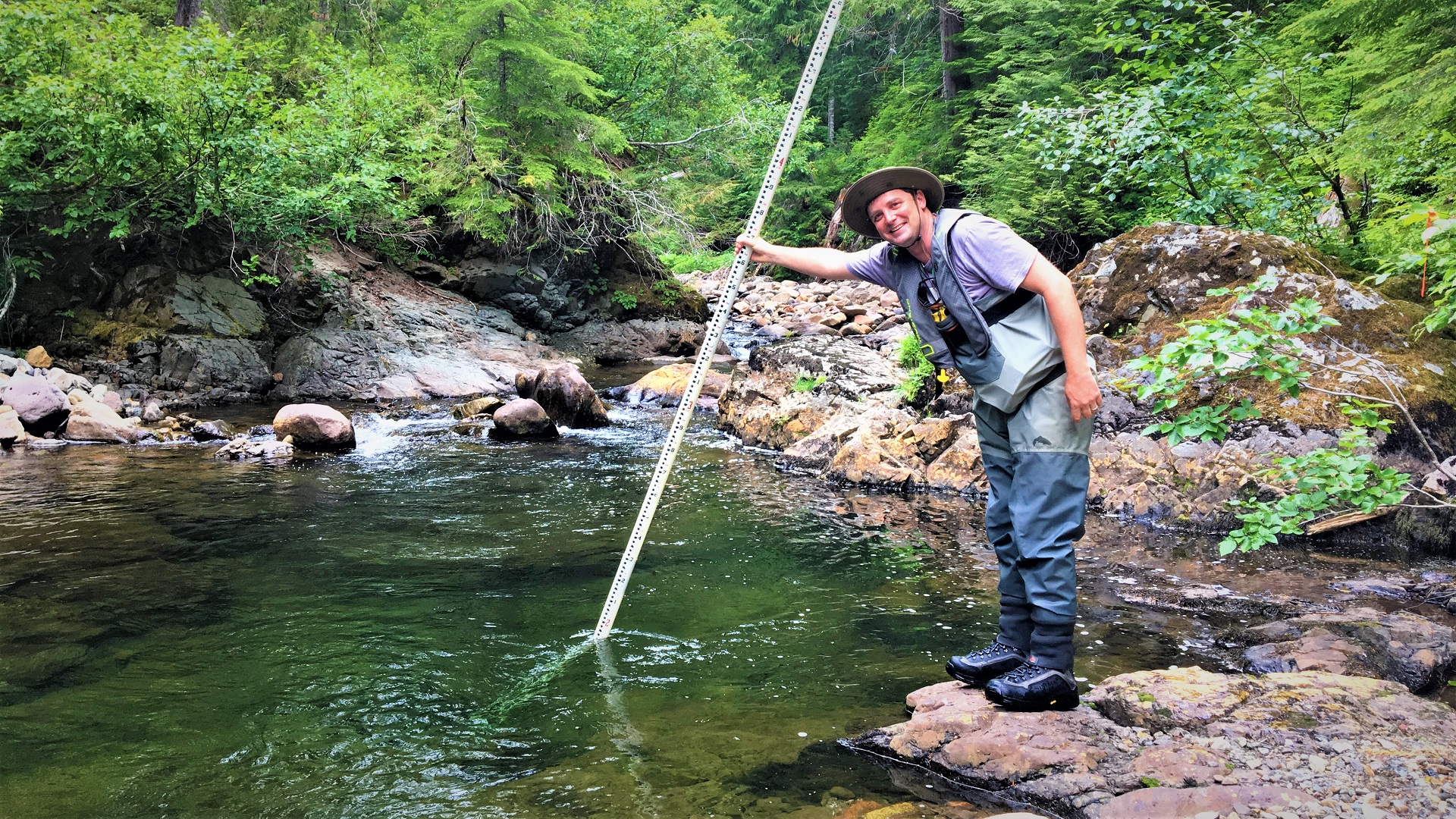 A person leaning over a pool, standing on a rock with a measuring rod.