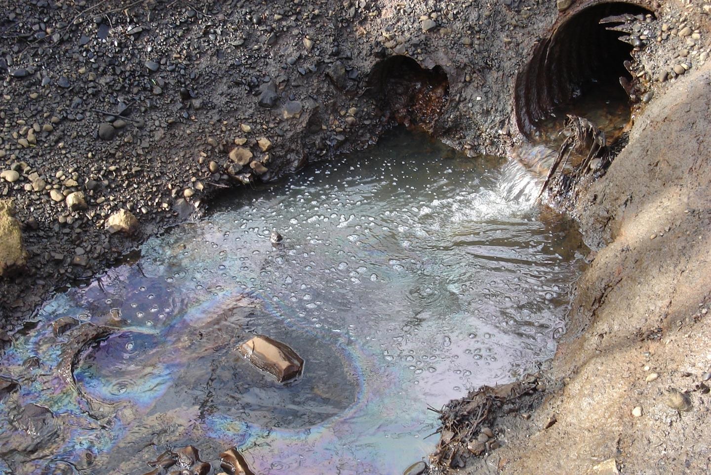 Oily water, with rainbow sheen on the surface, flows out of a culvert. into a ditch.