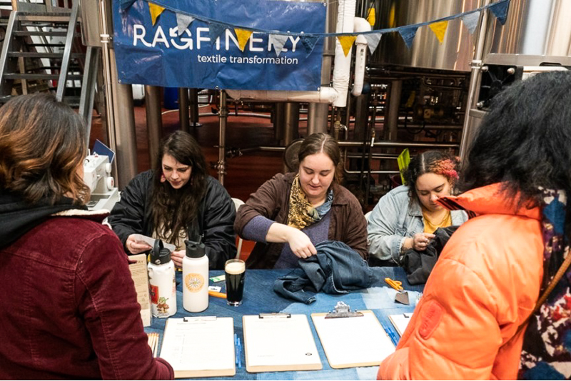 a group of women sitting behind a table, repairing clothing while others watch them.