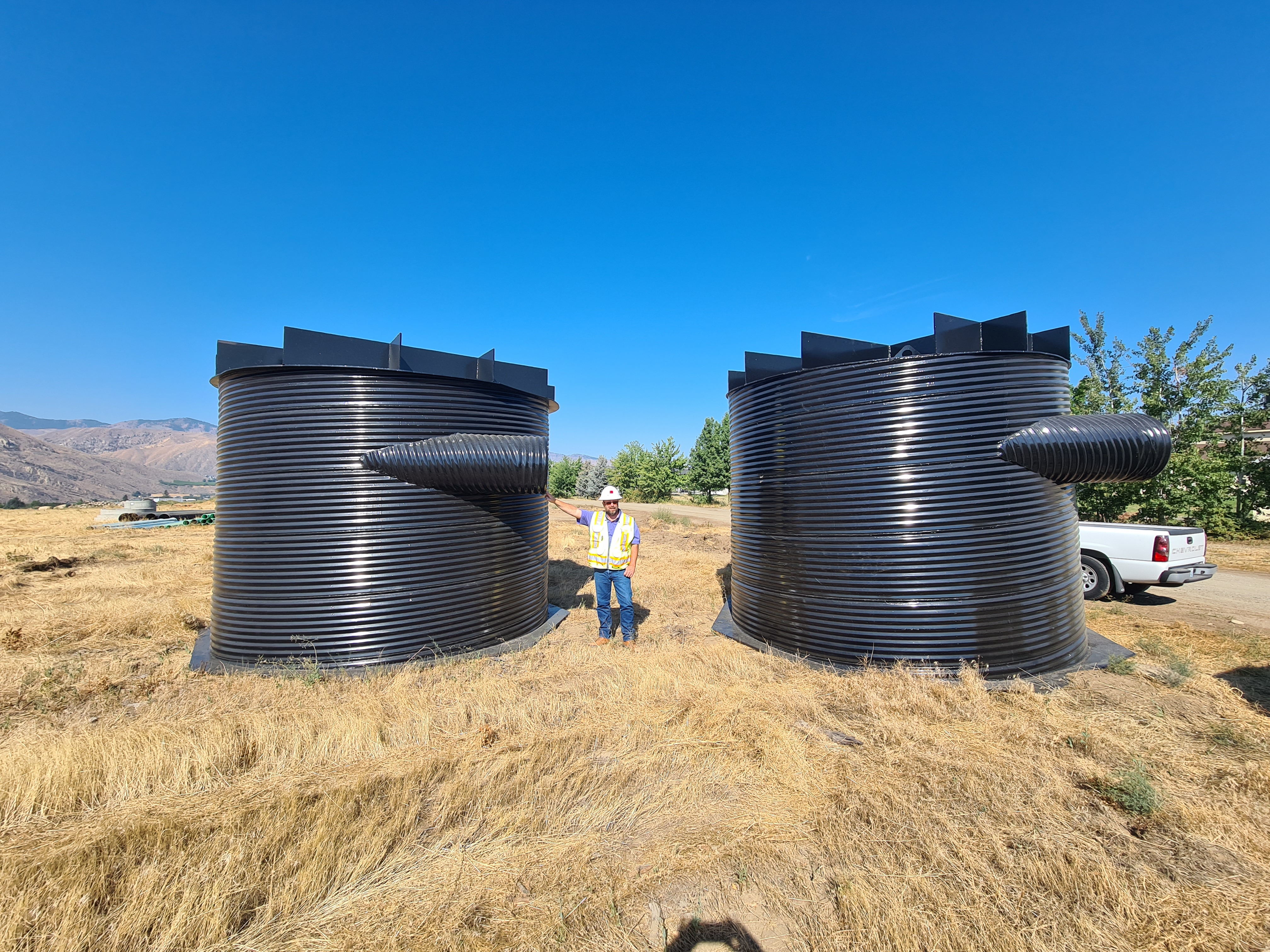 person in hard hat and safety vest standing next to very large cylinders. Person only is half as high as cylinders. 