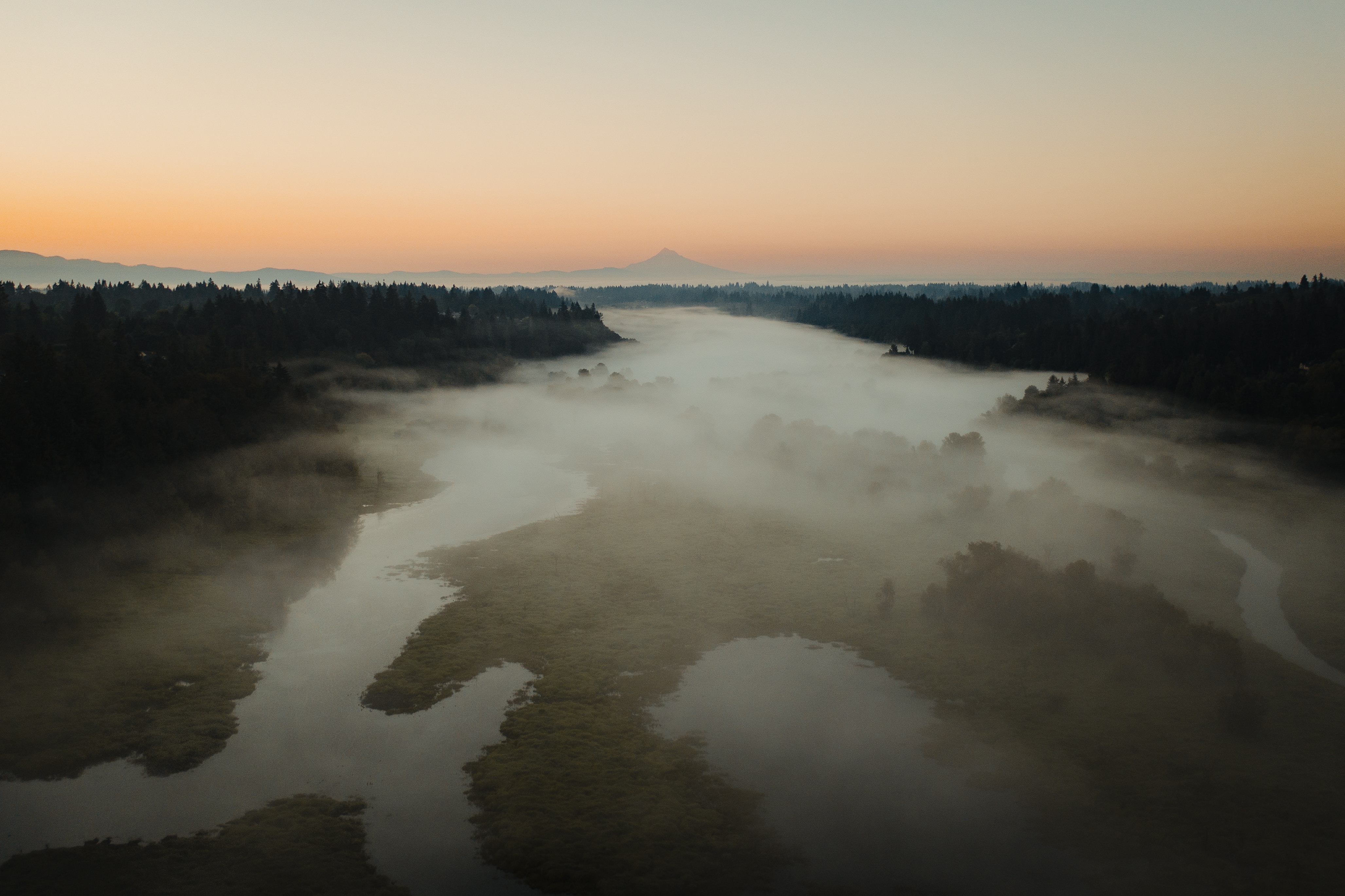 Aerial view of fog over a river with a sunset and Mount Hood in the distance.