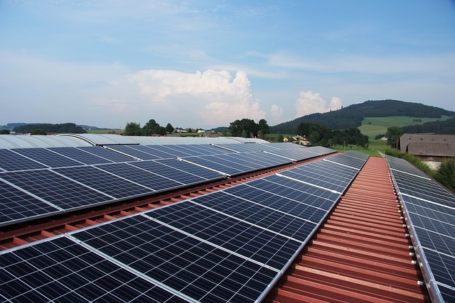 Three rows of solar panels atop a red roof, under a blue sky, and with a green forested hill in the background.