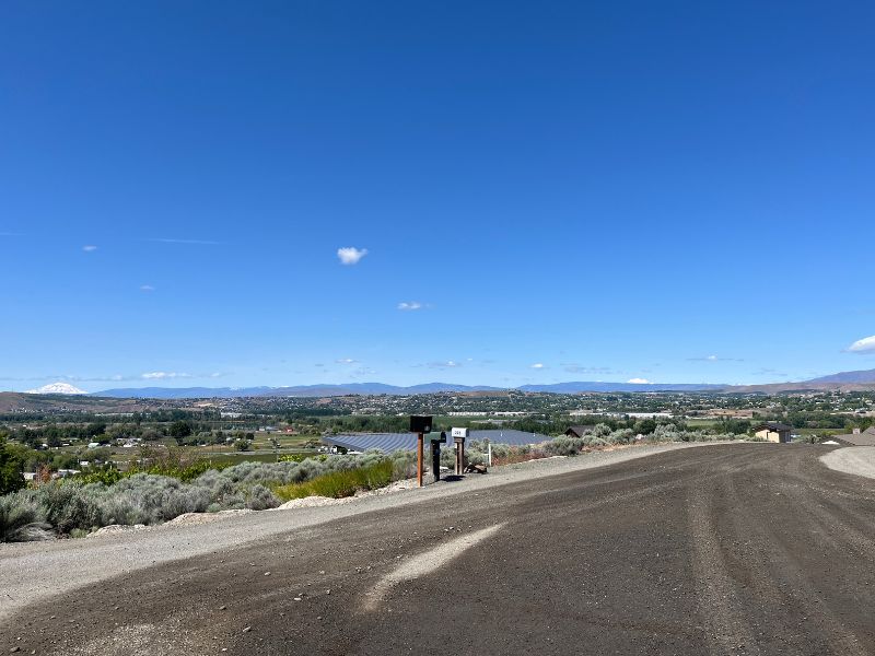 Una carretera de gravilla con tres buzones y vista de un valle con campos y edificios. Al fondo puede verse una cadena de montañas bajo un c