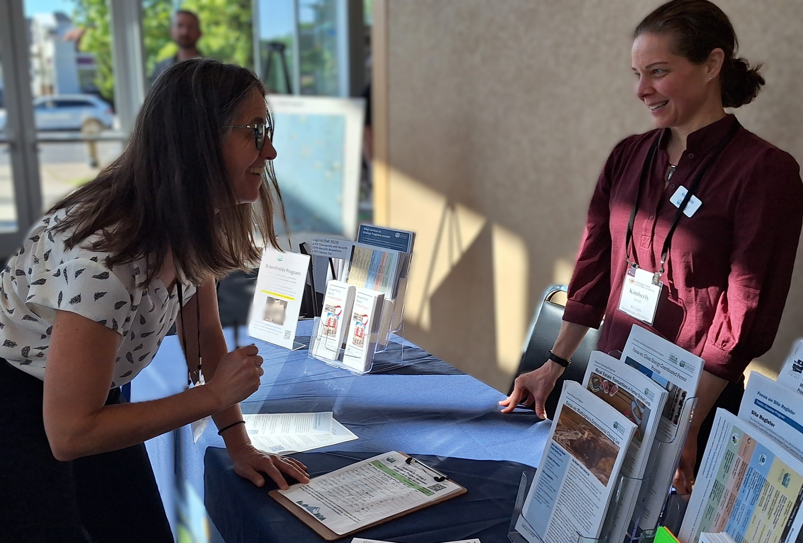 A smiling woman holds a pen over a clipboard on a table. A smiling female Ecology employee is on the other side of the table.