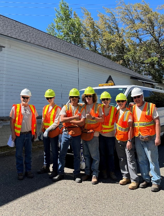 Group of young students wearing orange and yellow construction vests. Rod is standing with them, also in a construction vest.