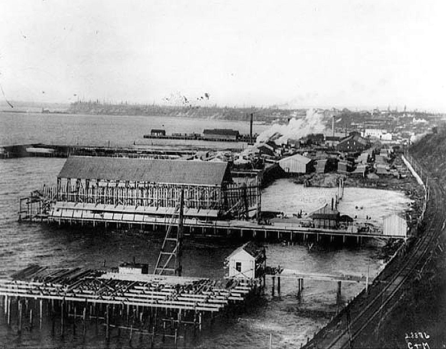Black-and-white photo of piers extending into water. A warehouse is on one pier, smoke is rising from the area behind, and train tracks run along the water.