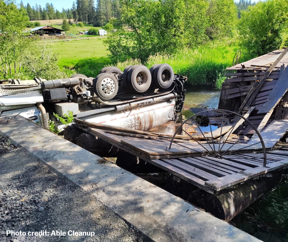 Overturned tanker truck lying on collapsed wooden bridge.