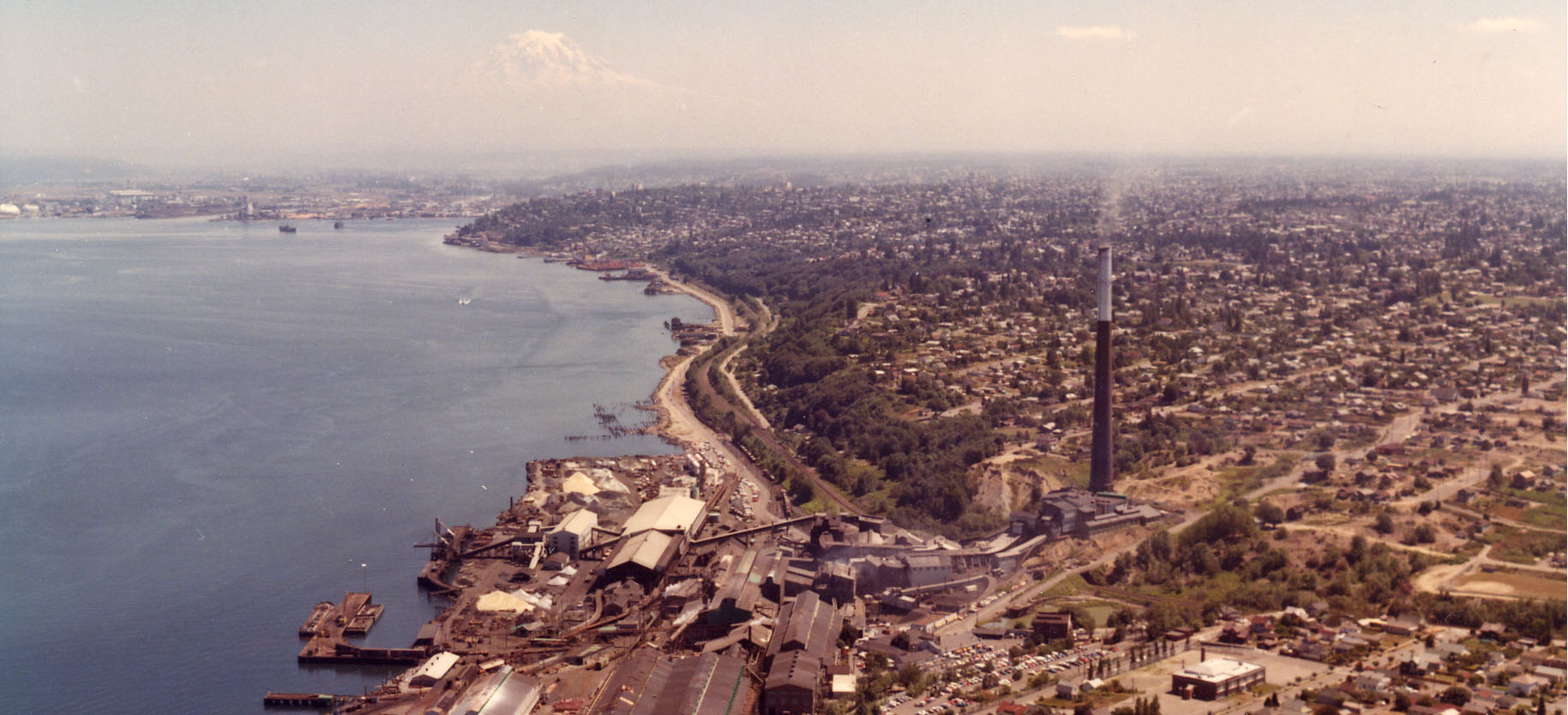 Old waterfront photo, mountain in background, tall industrial smokestack in foreground.