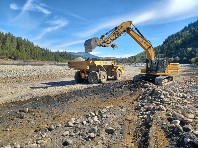 A backhoe excavates slag from the public beach and deposits it in a dump truck.