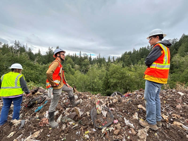 Three workers in orange vests and hard hats stand on top of a large garbage heap that looks over the tops of green trees in the distance.