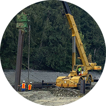 Excavator-run pile driver in gravel and sand with two workers.