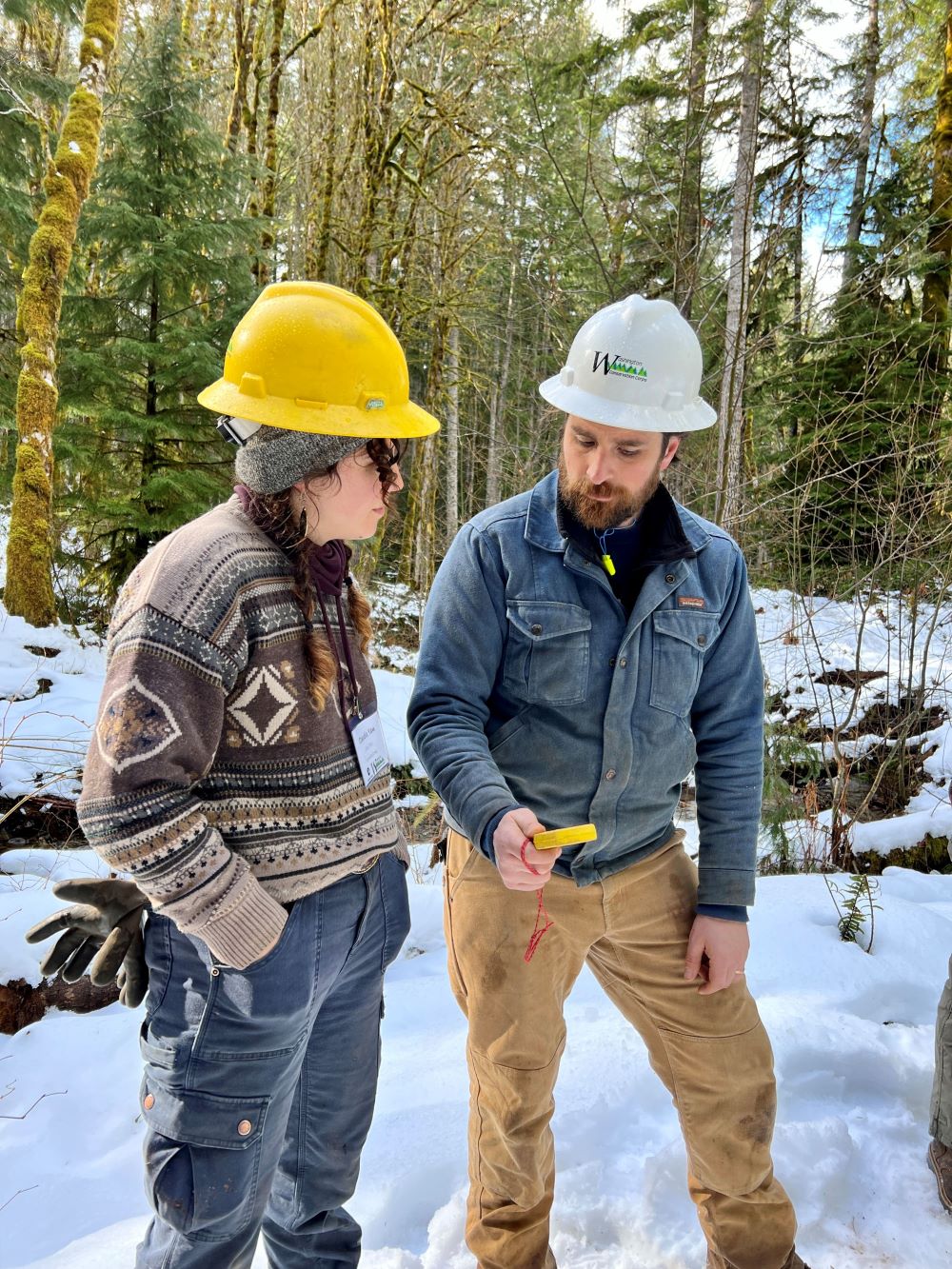 Josiah Downey stands with a WCC member on a snowy trail while teaching them how to use a clinometer.