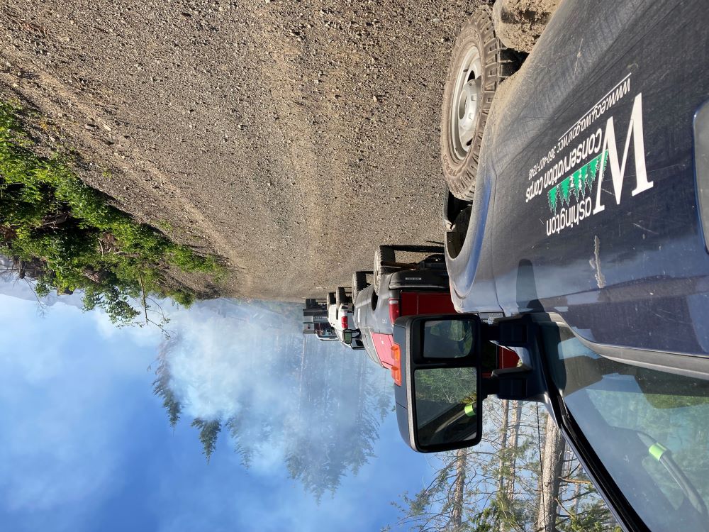 A view of a series of trucks driving up a gravel forest road toward a wall of wildfire smoke.