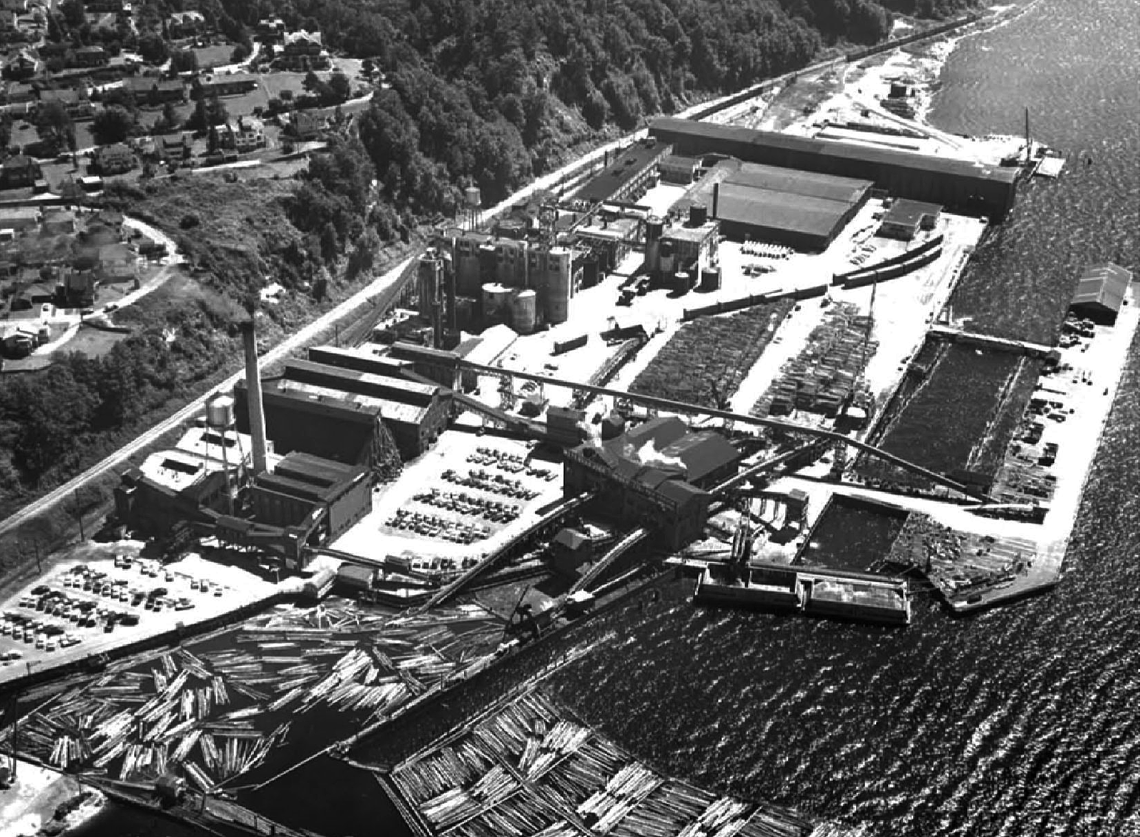 Industrial yard on waterfront with warehouses, smokestacks, and stacked logs.