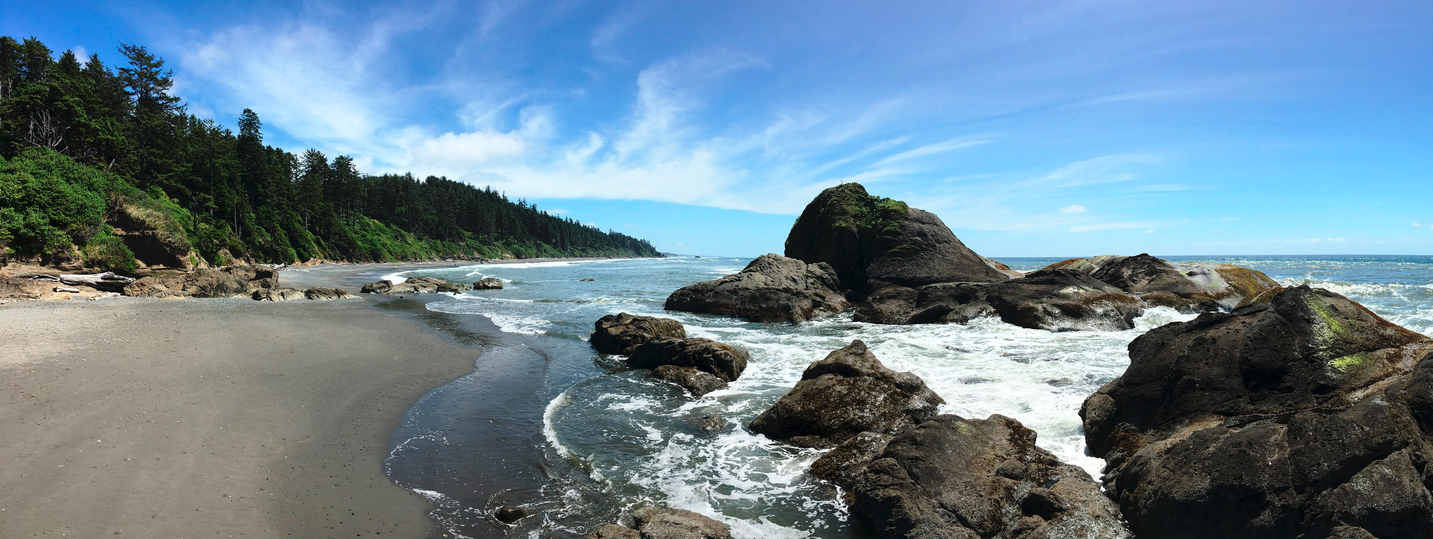Ocean coming in through large boulders onto a sandy beach on a beautiful blue sky day.