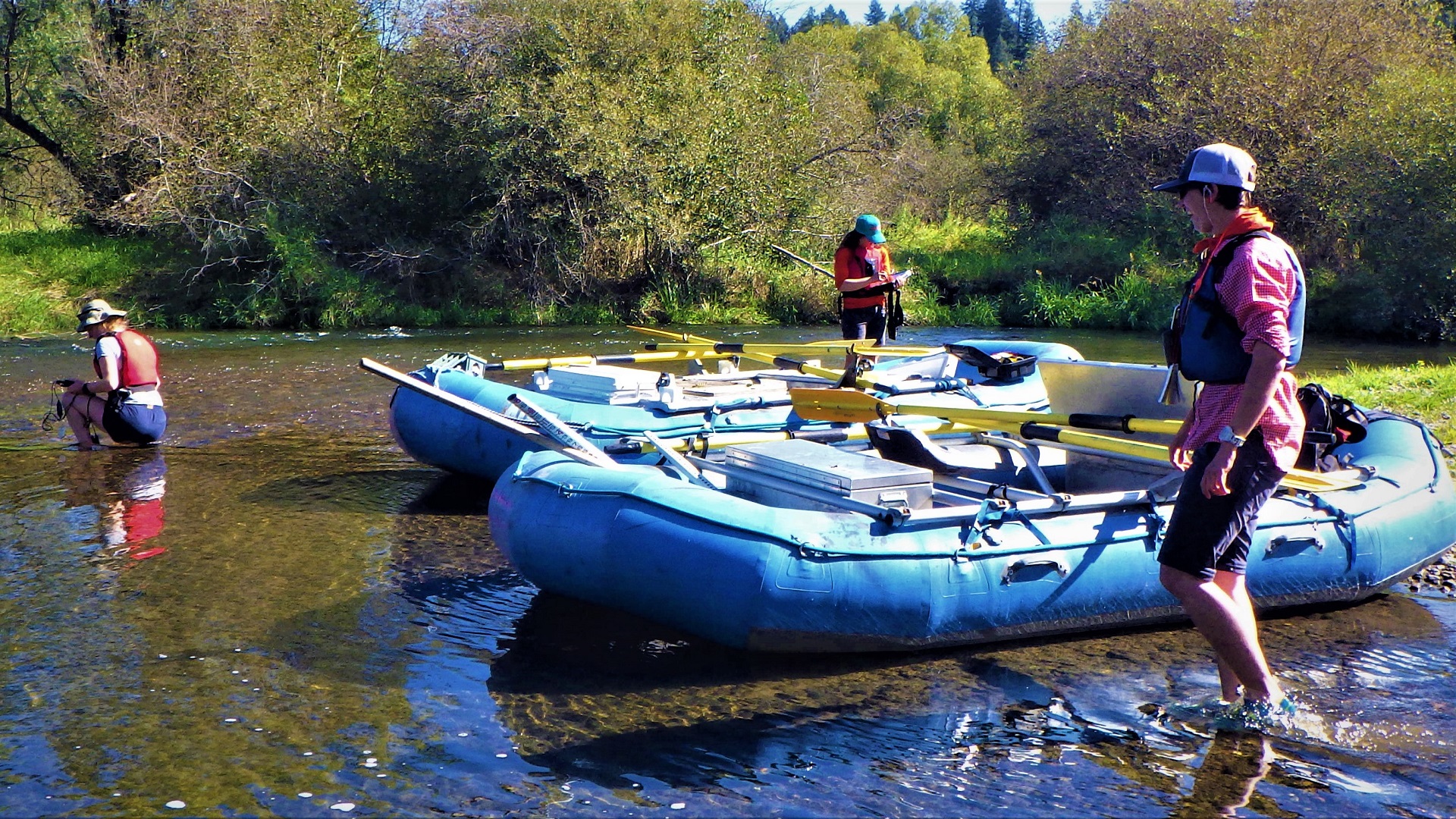 Crew members and two large rafts pulled over to on stream bank.