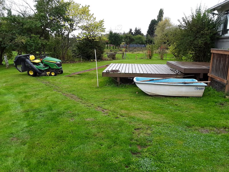 A grassy back yard with a small wooden deck, a small rowboat & small green tractor on the lawn