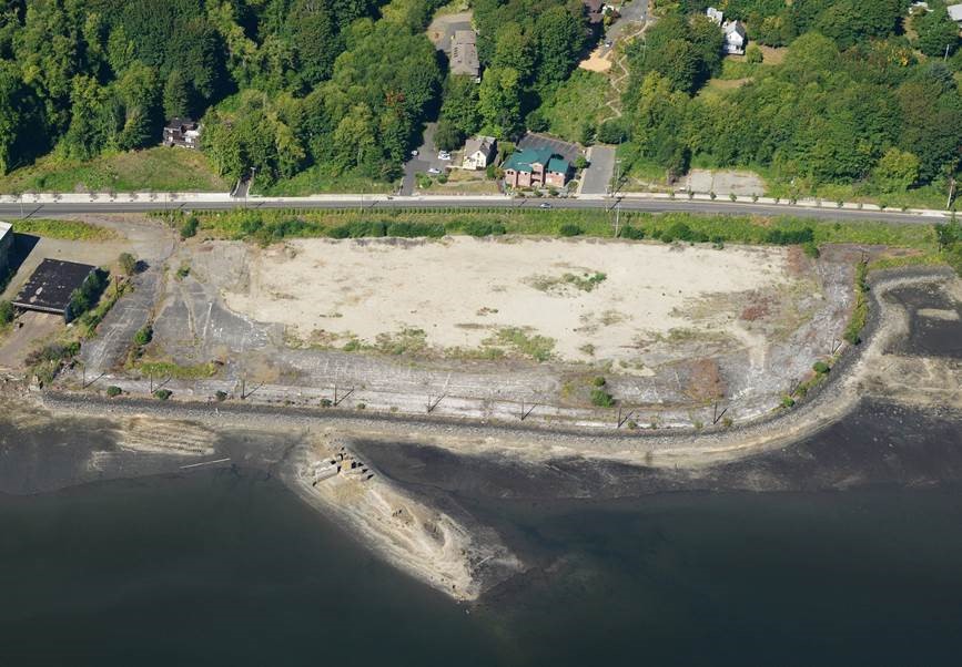 An aerial view of a shoreline with bare ground up against the water and trees in the background.