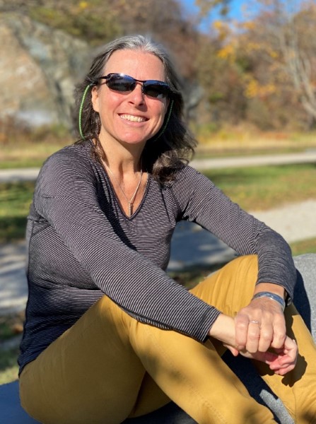 Rebecca Benjamin sits on a grassy field and smiles at the camera. She is wearing sunglasses and a long sleeved shirt.