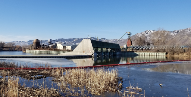 Water behind Zosel Dam. with grass in the foreground and snow-capped hills in the background.