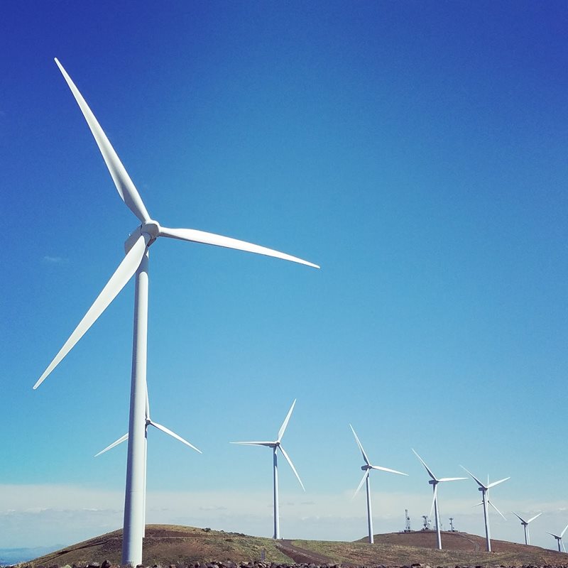 A large wind turbine is in the foreground against a cloudless blue sky. In the distance, several other wind turbines stand along a ridgeline