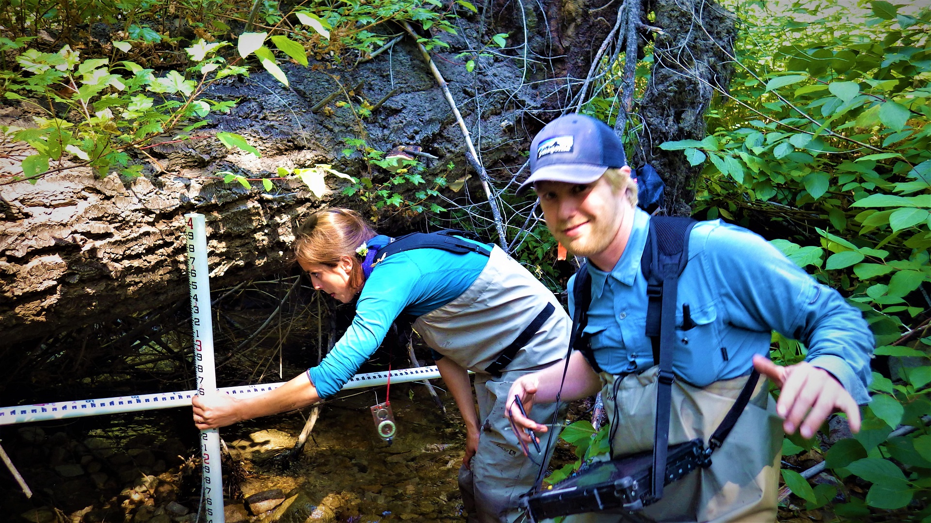 Two people stand in a stream holding measuring rods and a tablet