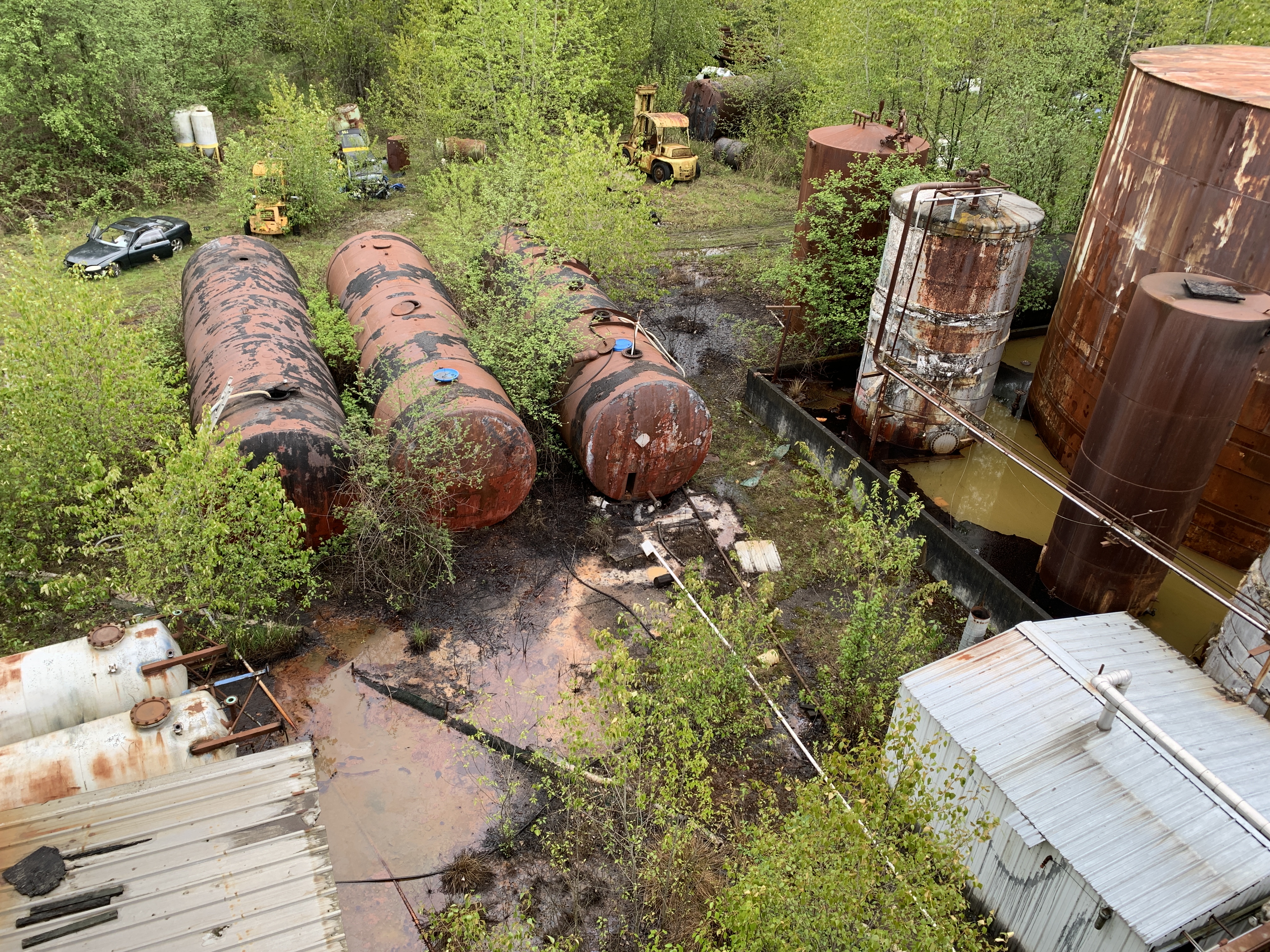 Looking down on large horizontal cylindrical tanks in center of photo, with large vertical tanks to the right. Liquid with rusty streaks has pooled underneath the tanks. 
