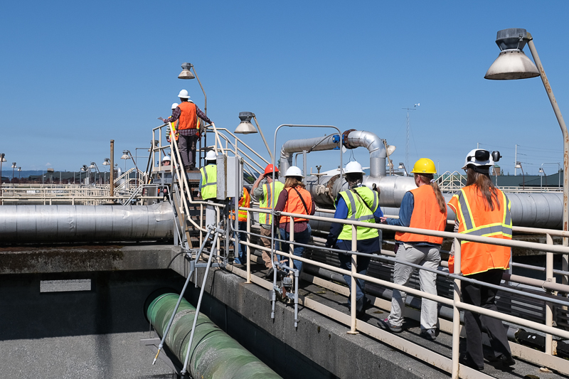 Several people wearing hard hats and safety vests cross past wastewater tanks on an elevated walkway.