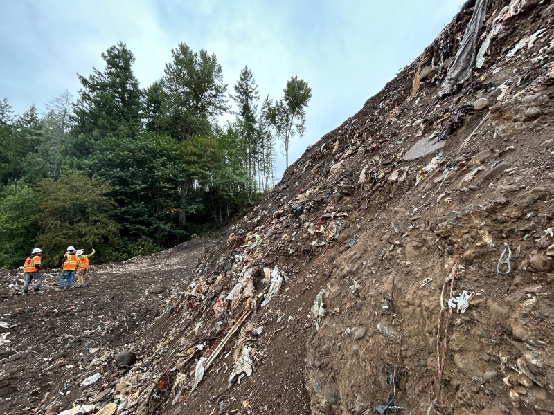A large mound of garbage and dirty soil towers over three small cleanup workers in orange vests and hard hats.