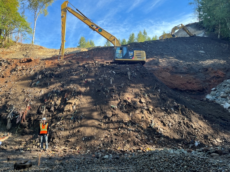 A worker in an orange vest and white hard hat stands at the base of a steep garbage slope whlie a large piece of machinery called an excavator with a long scoop shovel arm rests above him