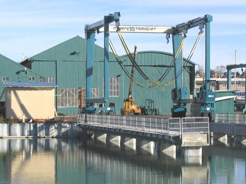 A blue boat lift at a dock in front of some mallard green industrial buildings.
