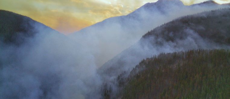 Wildfire smoke rising off of rolling hillsides under an orange sky 