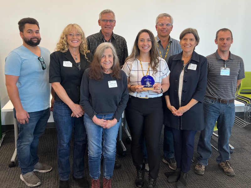 Eight people smile for the camera. Person in center holds award.