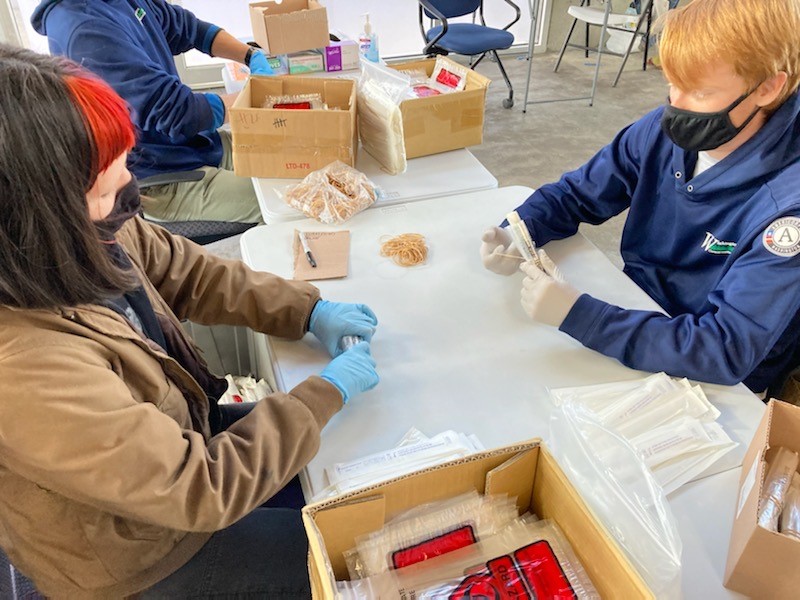 Three WCC members sit around a plastic folding table and place rubber bands around Covid-19 test kits.