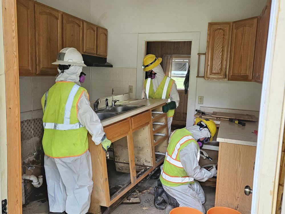 Two WCC members lift a cabinet and move it out of a kitchen. Another member look for water damage in a nearby cabiner.